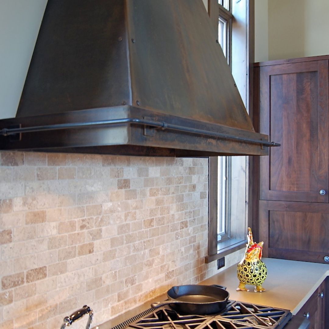 Breckenridge gallery1 features a kitchen stovetop with a large dark metal range hood, beige brick backsplash, black frying pan, decorative ceramic rooster container on the counter, and dark wooden cabinets in the background.