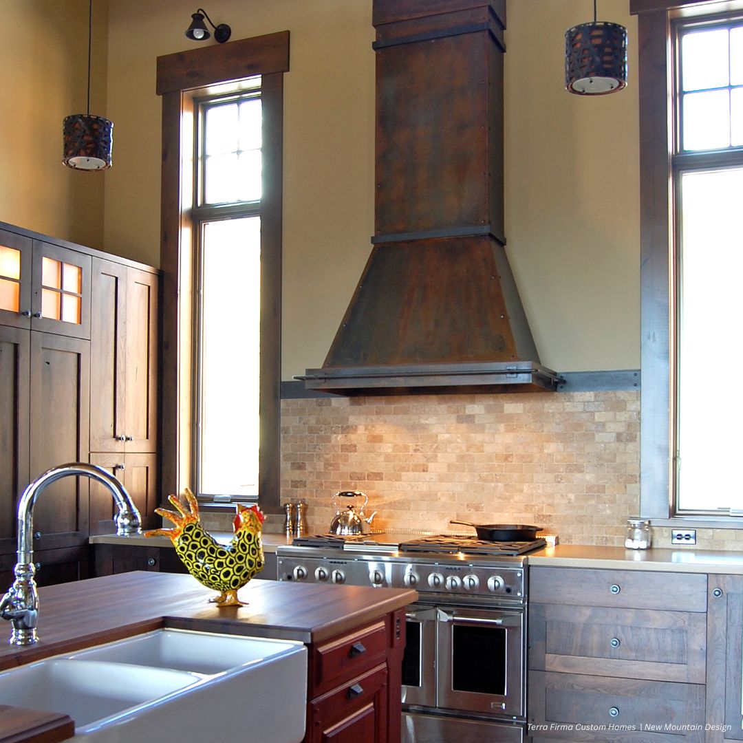 The Breckenridge gallery2 features a cozy kitchen with wooden cabinets, a farmhouse sink, a metal range hood, and a rooster-shaped teapot. Sunlight pours through tall windows, illuminating the inviting space.