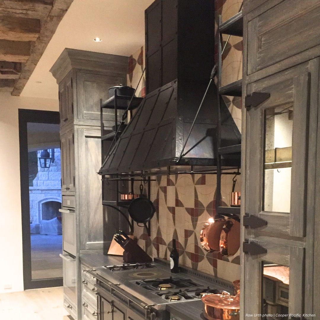A rustic kitchen with Breckenridge gallery3 gray wooden cabinets, a large black metal range hood, hanging copper pots and pans, a patterned backsplash, exposed wooden ceiling beams, and a stone fireplace in the next room.