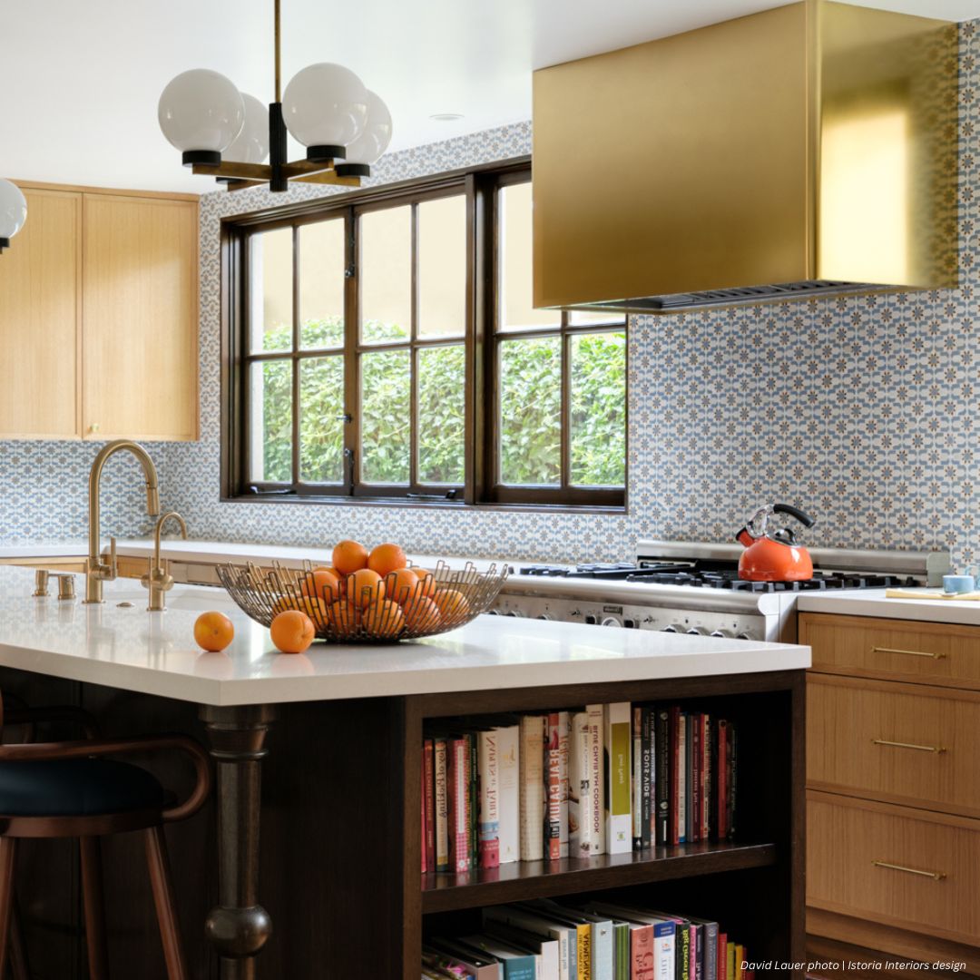 The Broadmoor gallery1 kitchen showcases a patterned tile backsplash, gold range hood, light wood cabinets, and a spacious island with bookshelves, white countertop, brass faucet, fruit bowl of oranges, and spherical pendant lights above.