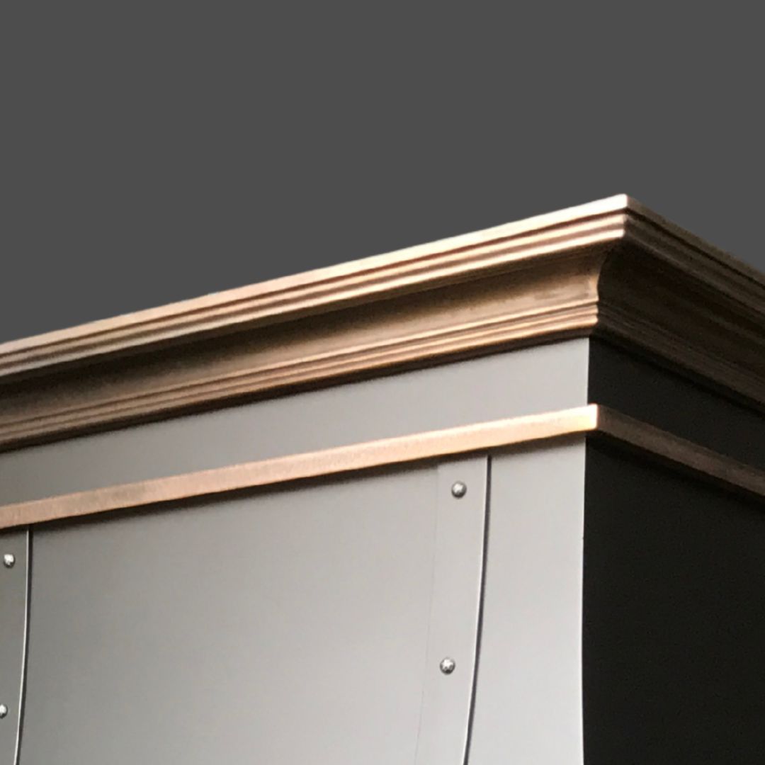 Close-up of the upper corner of the Bronze Cast 3 gray cabinet, featuring metallic trim and decorative molding, set against a plain dark background.