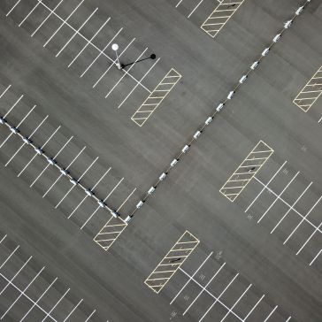 Brushed 5 Aerial view of Brushed 5: an empty parking lot with neatly lined spaces, yellow diagonal no-parking zones, a dividing barrier, and a single tall light pole casting a shadow.