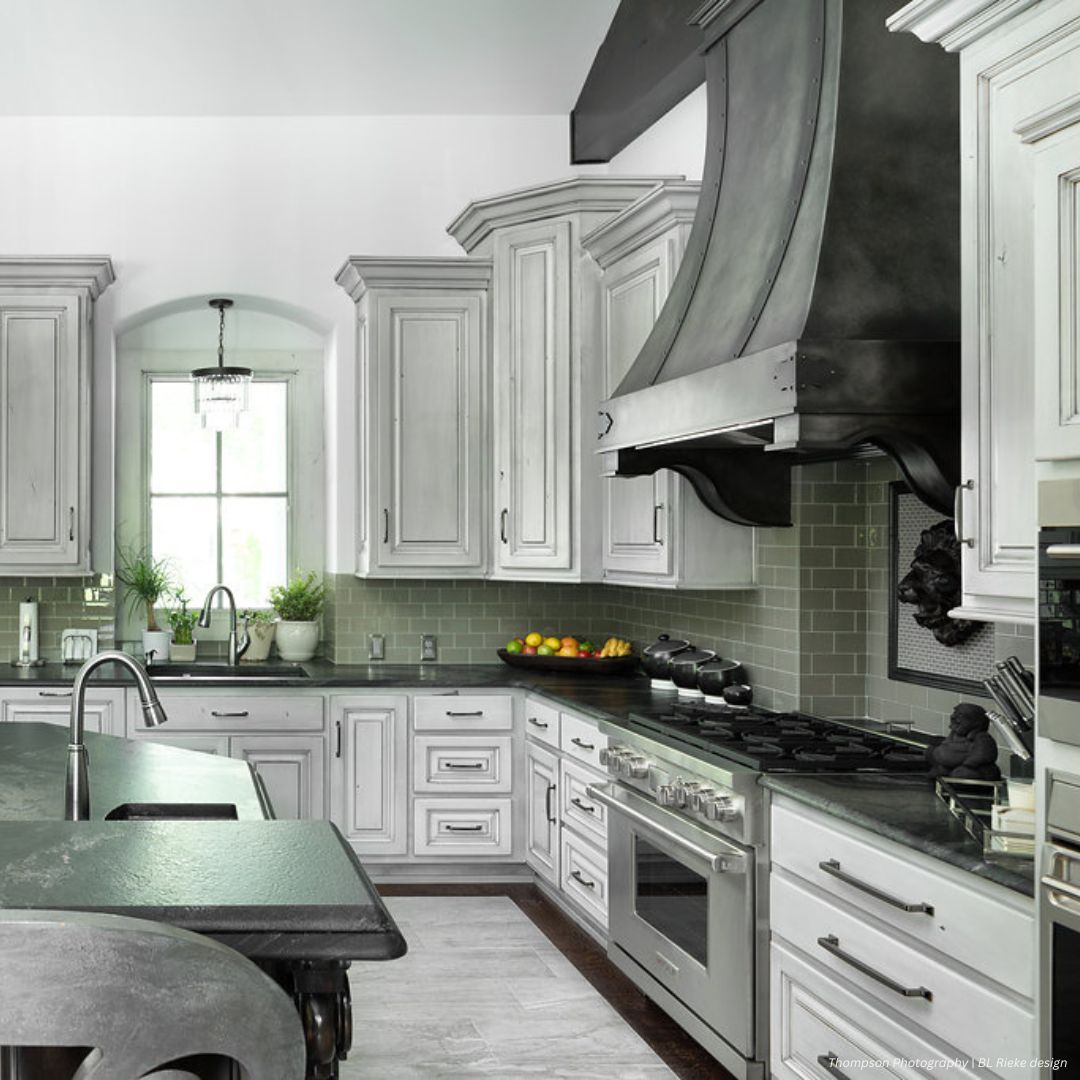 Cherry Hills gallery2 The Cherry Hills gallery2 features a modern kitchen with light gray cabinets, a bold dark range hood, stainless appliances, dark countertops, natural light from a window, and colorful accents from potted plants and fruit bowls.