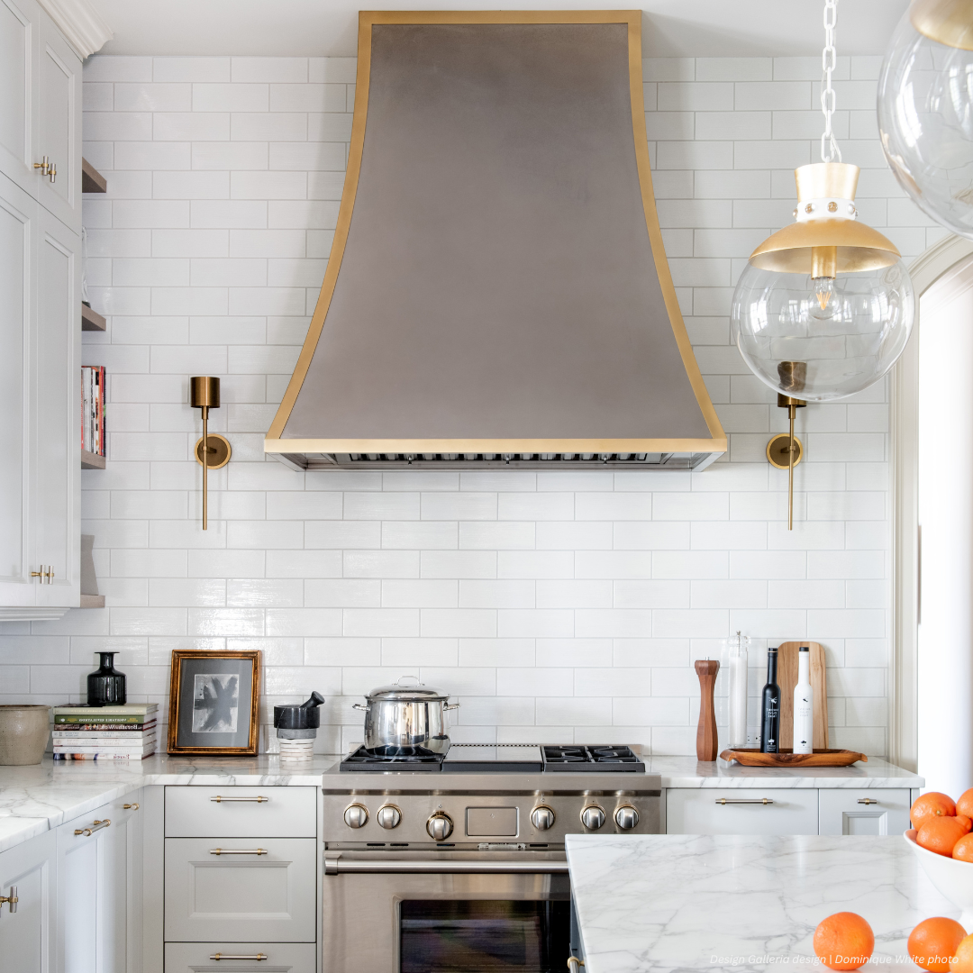 Dove Creek gallery1✅ The Dove Creek gallery1 features a modern kitchen with white cabinets, marble countertop, stainless steel stove, metallic range hood with gold trim, glass pendant lights, framed art, and oranges in a bowl.