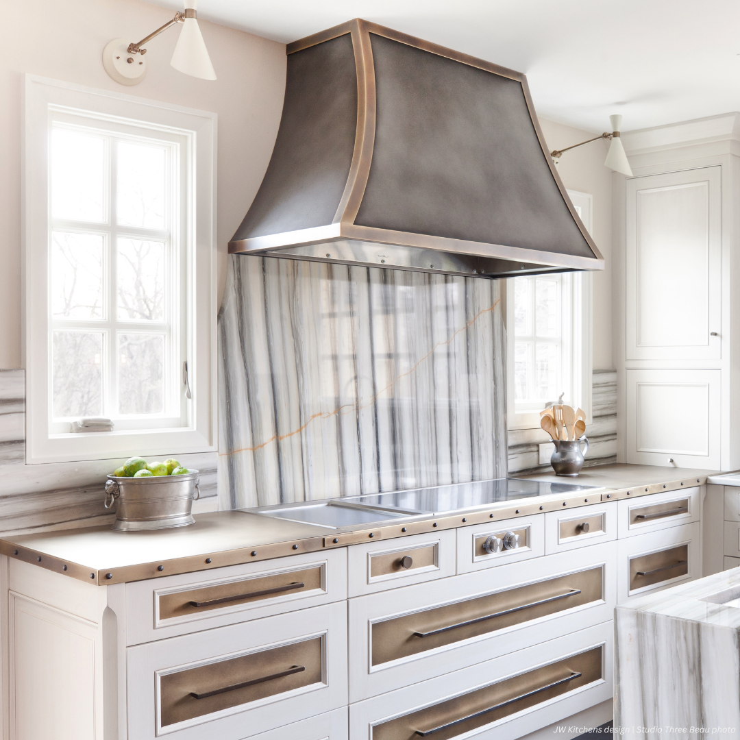 Dove Creek gallery2 The Dove Creek gallery2 showcases a bright kitchen with marble backsplash and counters, brass-accented drawers, a large metal range hood, white cabinetry, two windows, and a bowl of green apples on the countertop.