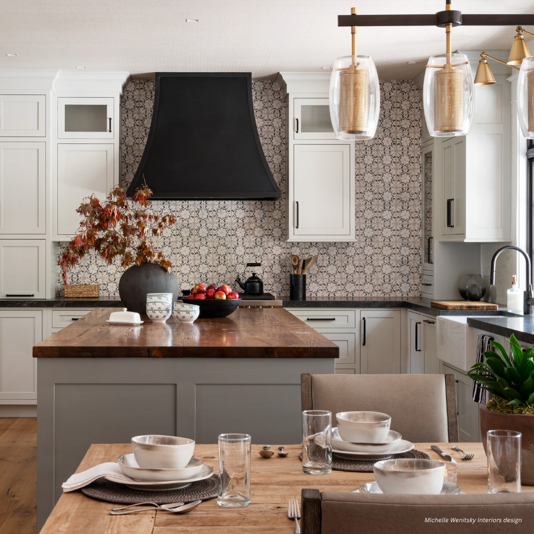 Dove Creek gallery3 The Dove Creek gallery3 kitchen features white cabinets, a patterned tile backsplash, a bold black range hood, a wooden island countertop, and a dining table adorned with dishes, glassware, and neutral-toned decor.