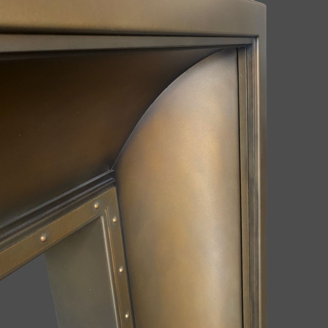 Close-up of the Foundry 3-2’s metallic corner featuring a curved, smooth bronze or dark brass finish beside a riveted panel, set against a solid gray background.