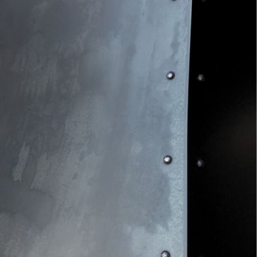 A close-up of Gunmetal 1, showing its curved edge and several evenly spaced rivets against a black background. The metallic surface appears slightly weathered and textured.