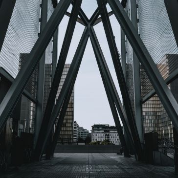 View through a modern structure featuring Gunmetal 3, with large metal beams forming triangles, looking out at city buildings beneath a cloudy sky.