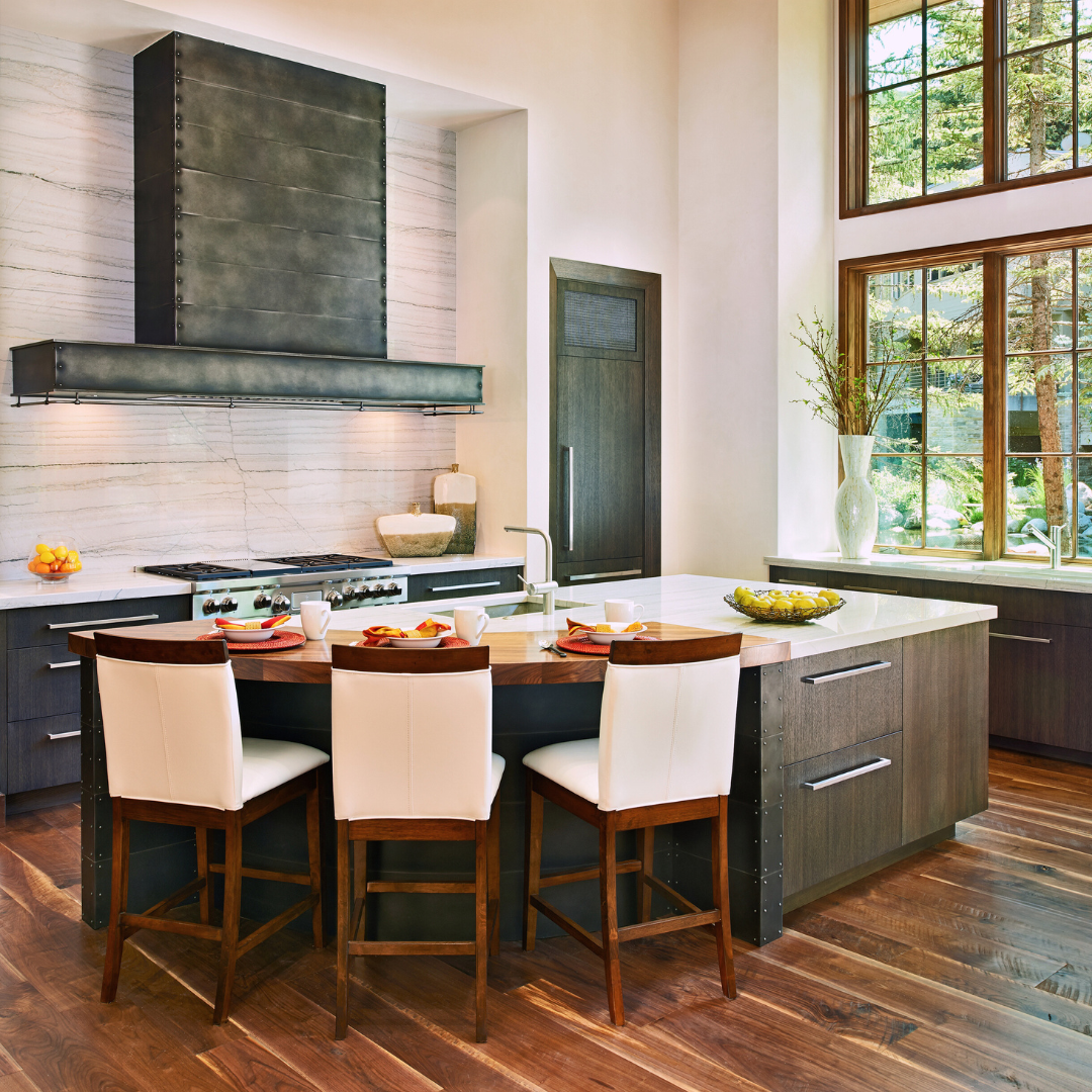 The Leadville gallery2✅ kitchen features stainless steel appliances, a marble backsplash, dark wood cabinets, a spacious island with sink, three white barstools, and large windows offering natural light and tree views.