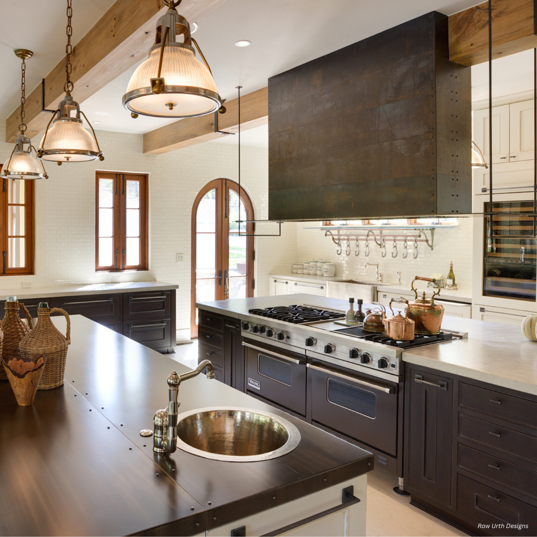 Leadville gallery3✅ (1) features a spacious, modern kitchen with dark cabinets, a large island with sink, stainless steel stove, built-in shelves, pendant lights, wood ceiling beams, and sunlight from arched windows.