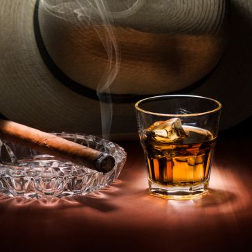 Leathered 7032 A glass of Leathered 7032 whiskey with ice stands by a lit cigar in a crystal ashtray. In the background, a large hat adds to the refined and relaxed ambiance.