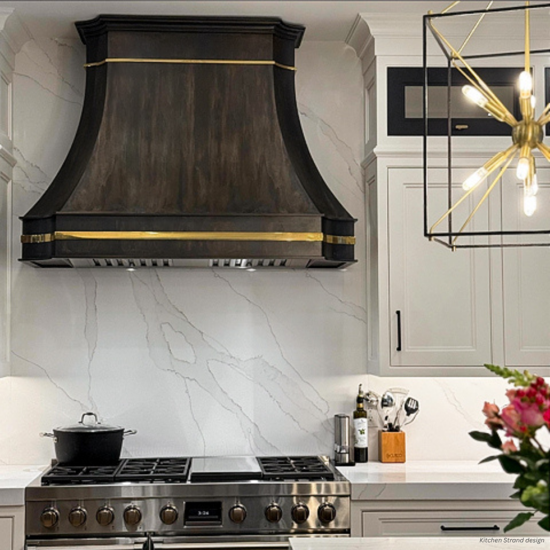 Mary Jane gallery1 A modern kitchen featuring the Mary Jane gallery1—a large black and gold range hood—marble backsplash, stainless steel stove, white cabinets, geometric chandelier, pot on the stove, utensils on the counter, and flowers in the foreground.