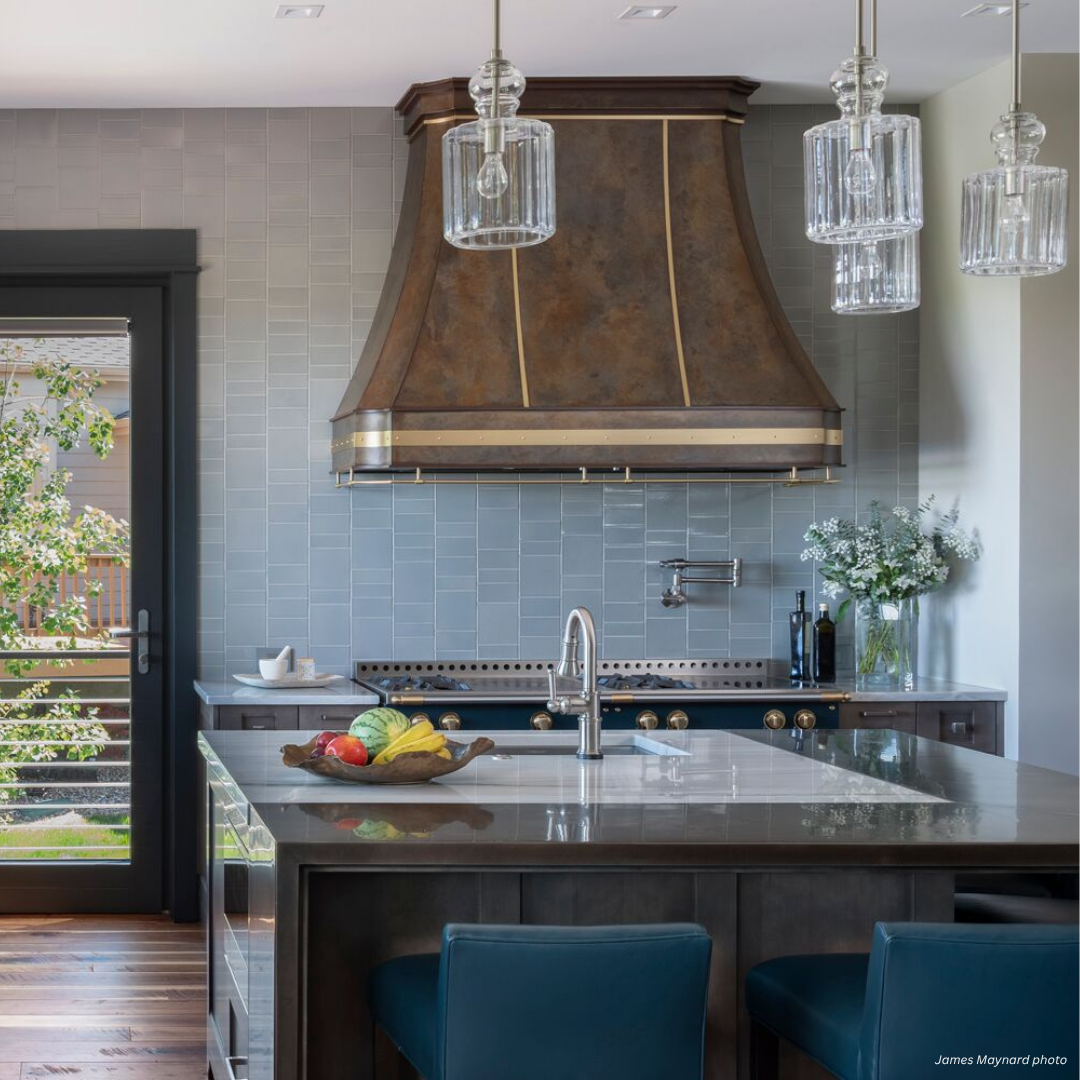 Mary Jane gallery2 The Mary Jane gallery2 kitchen features a spacious island, blue upholstered stools, pendant lighting, a metal range hood, gray tile backsplash, built-in stove, fresh produce on the counter, and an exterior door.