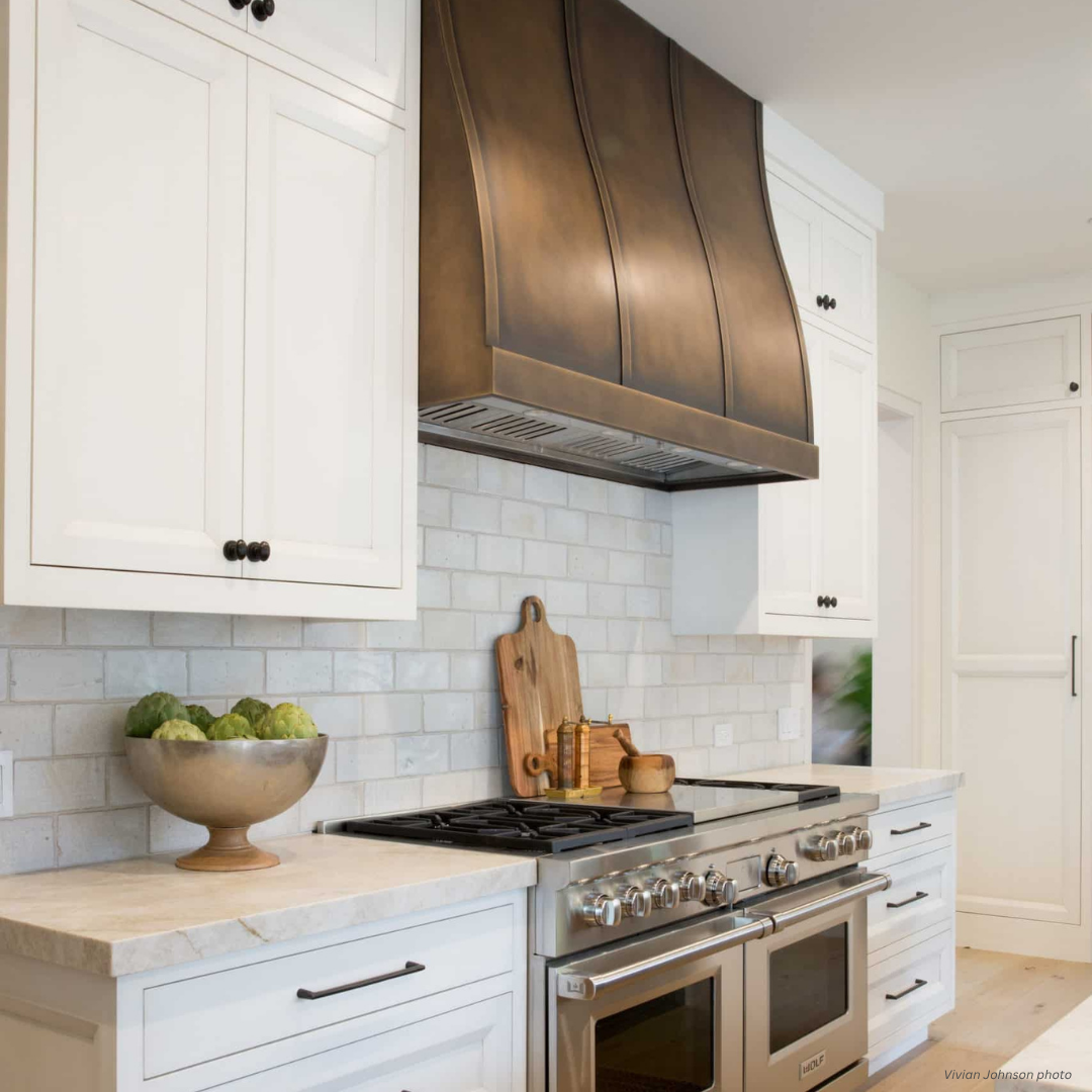 Masonville gallery3 The Masonville gallery3 features a modern kitchen with white cabinets, a large bronze range hood, subway tile backsplash, stainless steel stove, cutting boards, condiments, and a bowl of artichokes on the counter.
