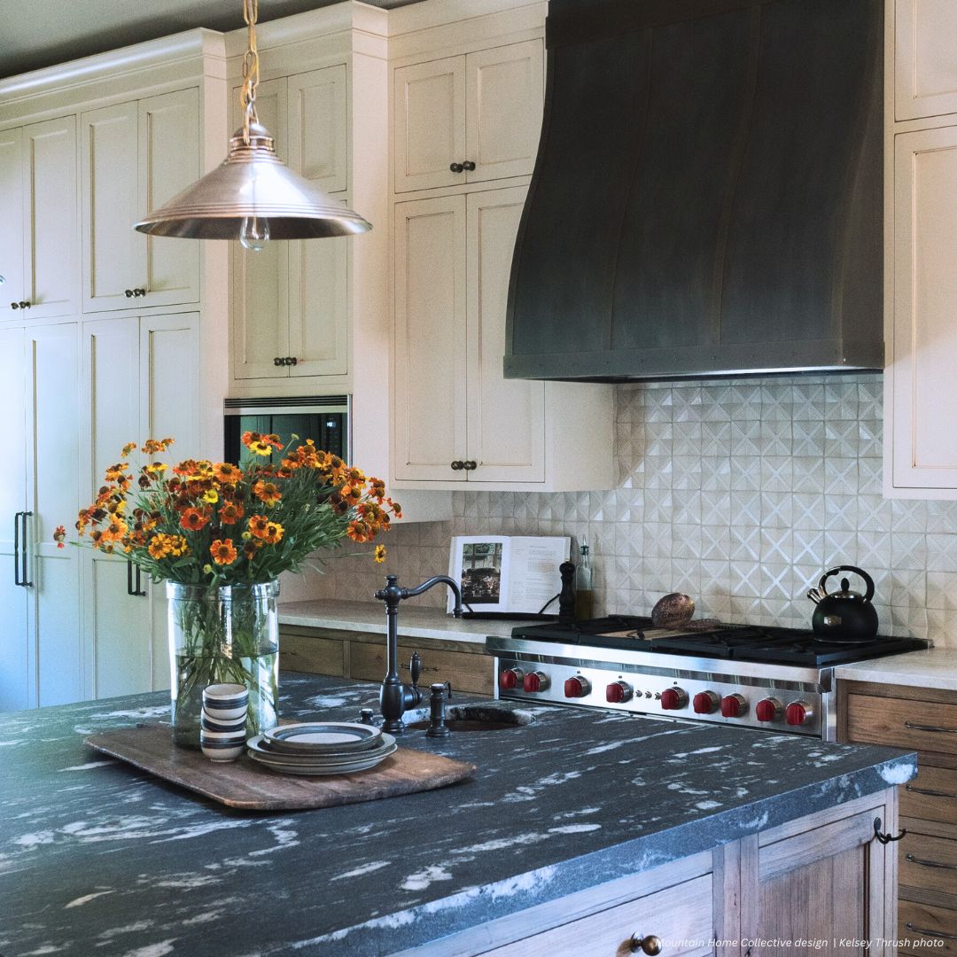 Masonville gallery3✅ The Masonville gallery3✅ kitchen showcases cream cabinets, a sleek metal range hood, a black kettle on the stove, and a marble island topped with flowers, plates, and cups arranged on a wooden tray.
