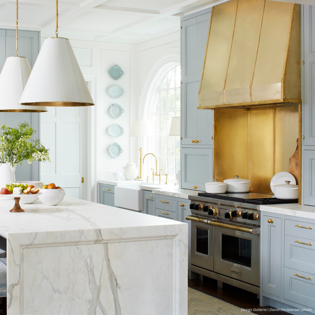 Mesa gallery2 The Mesa gallery2 kitchen features light blue cabinets, a spacious marble island, gold accents like a metallic range hood, white pendant lights, a window above the farmhouse sink, and decorative wall plates.
