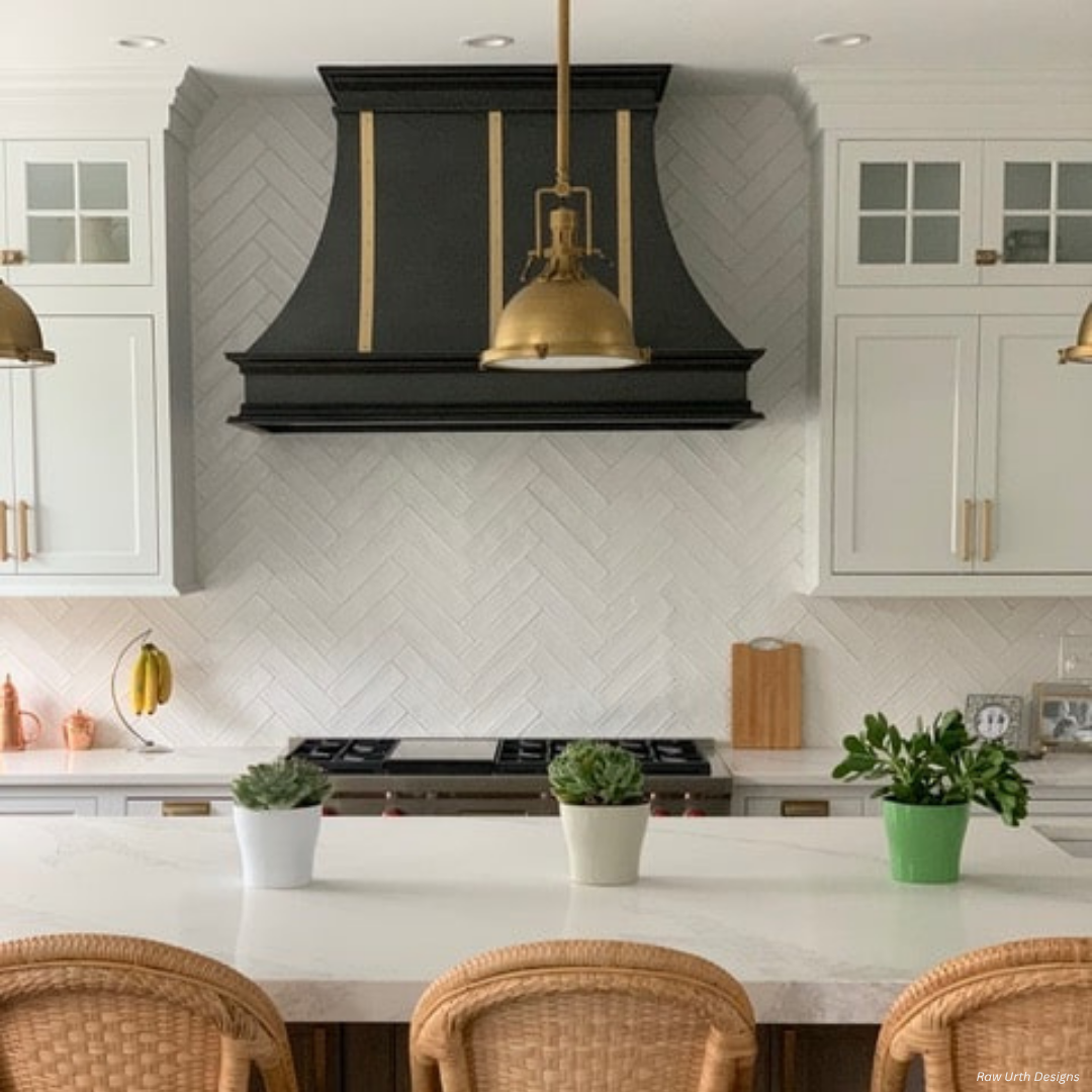Montrose gallery2 The Montrose gallery2 features a modern kitchen with a white herringbone tile backsplash, black and gold range hood, white cabinets, brass pendant lights, a white island, three potted plants, and wicker chairs.