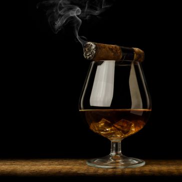 The Oil Rubbed 6 cigar rests on the rim of a snifter glass filled with amber liquor and ice cubes, set against a dark background.
