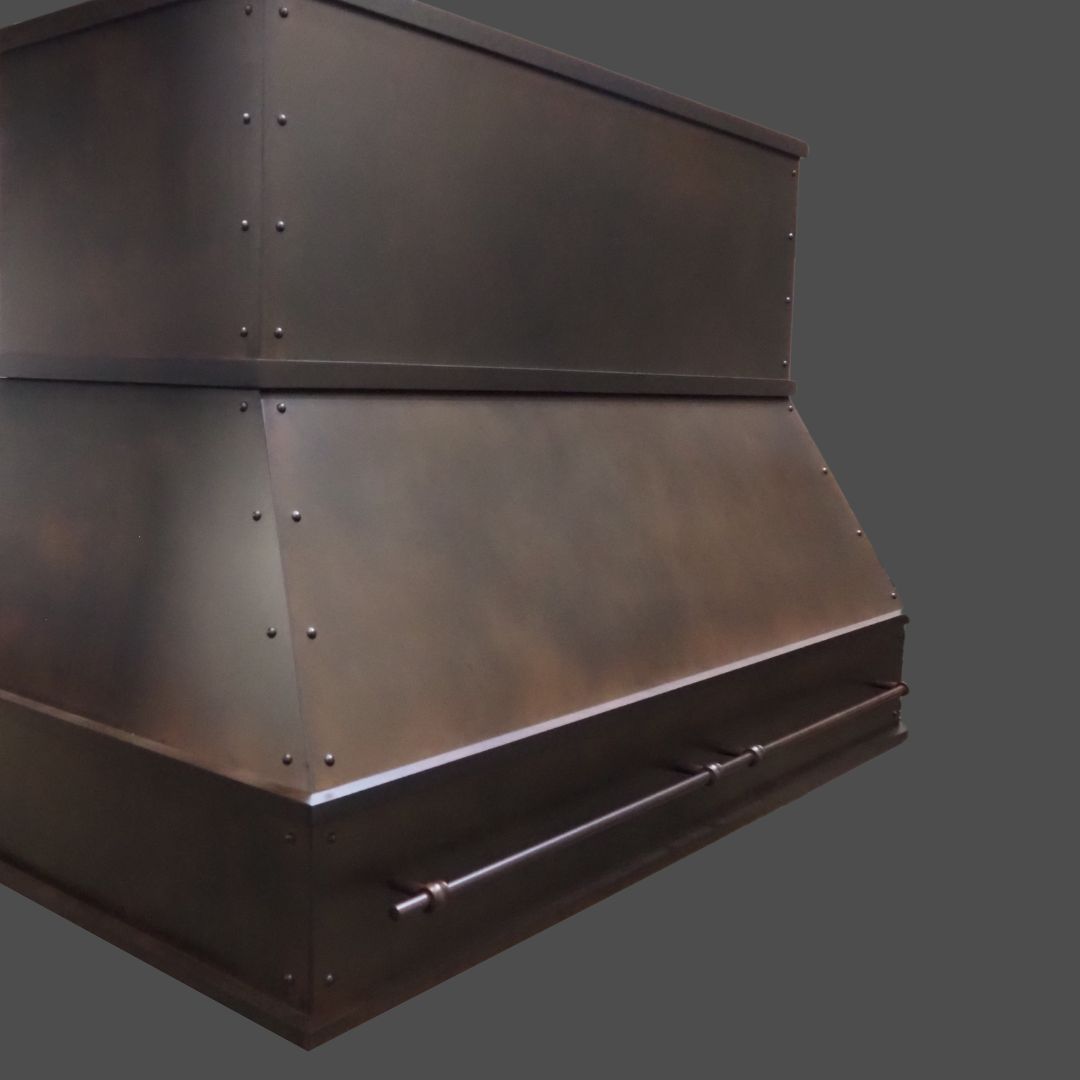 The Oil Rubbed Iron 3 is a large, dark metal range hood with a sloped midsection, edge rivets, and a horizontal bar at the bottom, displayed against a plain gray background.