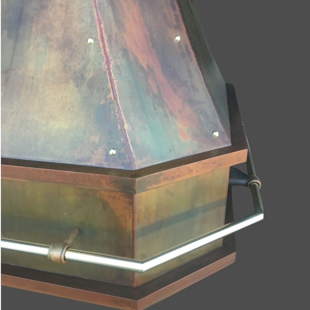 Penny Iron 2 Close-up of the Penny Iron 2 chimney hood, featuring a metallic hexagonal design with bronze and copper patina, metal rivets, and a silver handle, set against a plain gray background.