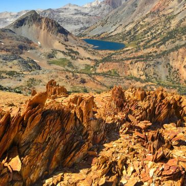 Penny Iron 6783 A scene featuring Penny Iron 6783: jagged orange rocks in the foreground, a mountainous landscape with green valleys, and a blue lake in the distance beneath a clear sky.