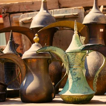 Penny Iron A set of Penny Iron Arabic coffee pots, featuring long spouts and pointed lids in mixed brass and greenish finishes, displayed outdoors under sunlight.