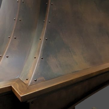 A close-up of Rustic Iron 1 showcases its curved, metallic surface with a bronze finish, visible rivets along the seams, and subtle color variations.