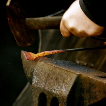 A close-up of Rustic Iron 6 being held with tongs on an anvil and struck with a hammer, sending sparks flying against a dark background.