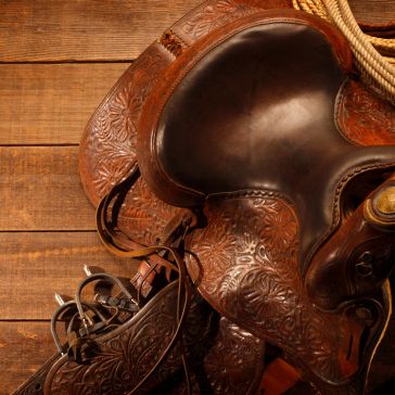 Rusty Rustic (2) Close-up of the Rusty Rustic (2) tooled leather western saddle with intricate floral patterns, resting on wood. A coiled rope and stirrup showcase this classic cowboy gear.