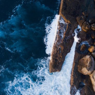 Slate 6893 Aerial view of waves crashing against rugged, brown Slate 6893 coastal rocks surrounded by deep blue ocean water.