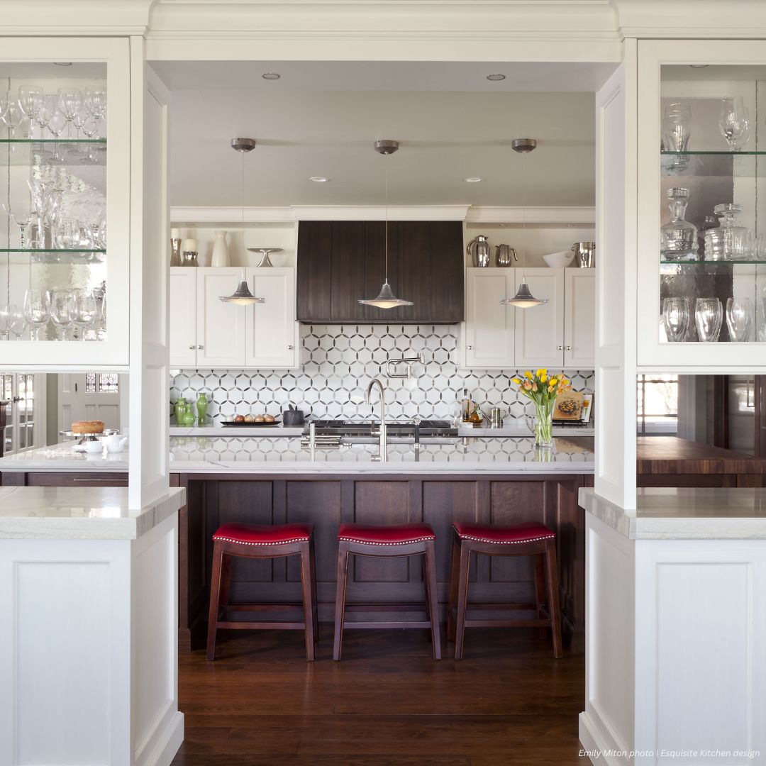 Snowmass Gallery3 The Snowmass Gallery3 features a modern kitchen with white cabinets, a dark wood island, red-cushioned stools, pendant lighting, hexagonal tile backsplash, glass shelves for glassware, and a vase of yellow flowers on the counter.