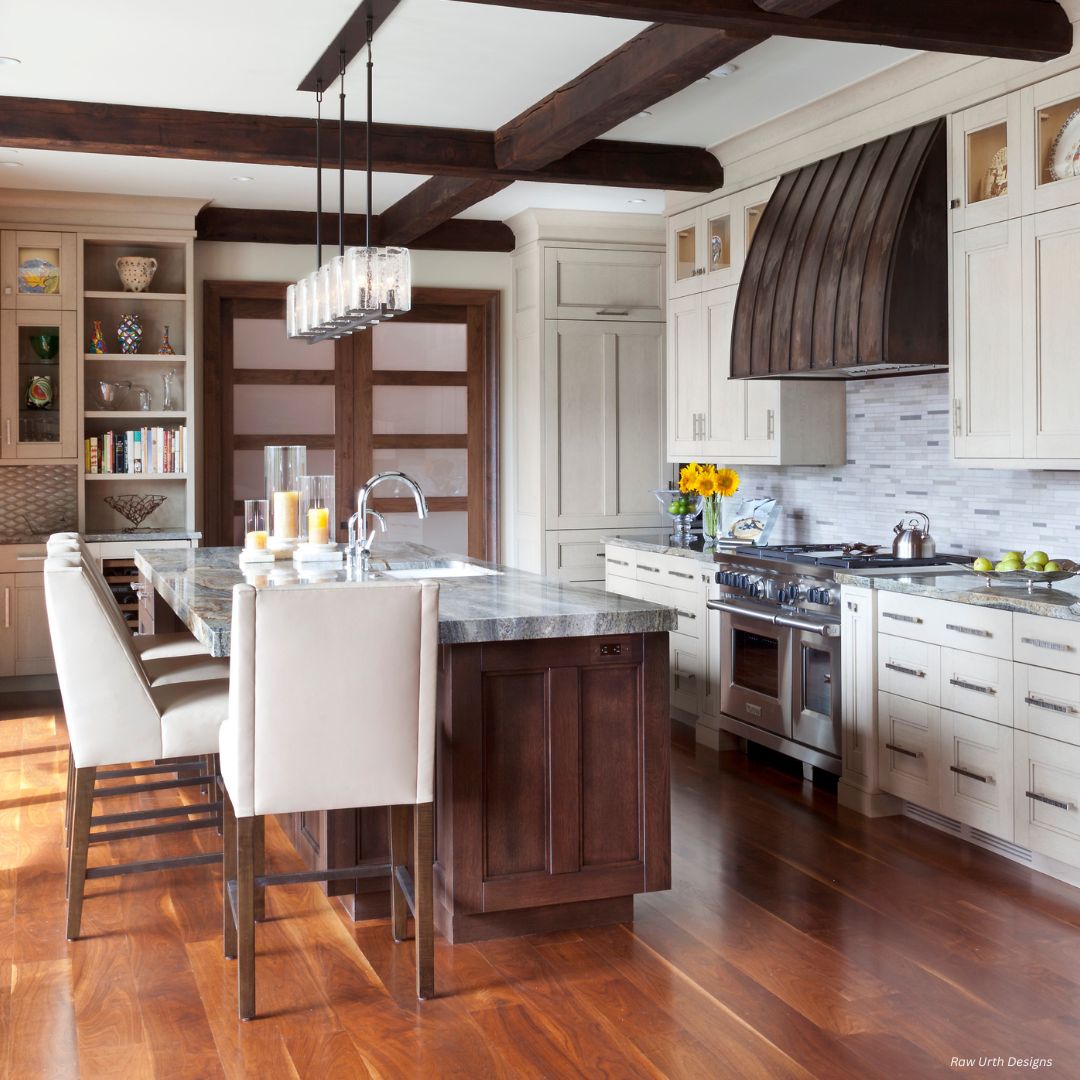 Snowmass gallery 2 The Snowmass Gallery 2 kitchen features a large island with marble countertop, white upholstered chairs, wooden floors, light cabinets, stainless steel appliances, pendant lights, and exposed wood beams. Sunflowers decorate the counter.