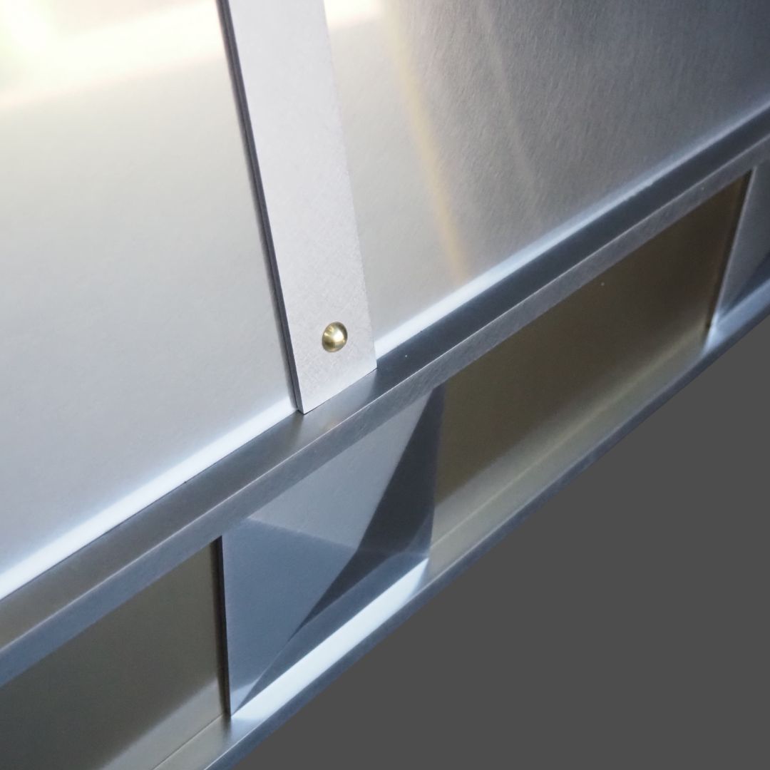 Close-up of Stainless Burnished 3 panels joined by a small brass screw, casting geometric shadows and reflections on silver-gray surfaces. The lower part of the image shows a dark gray background.