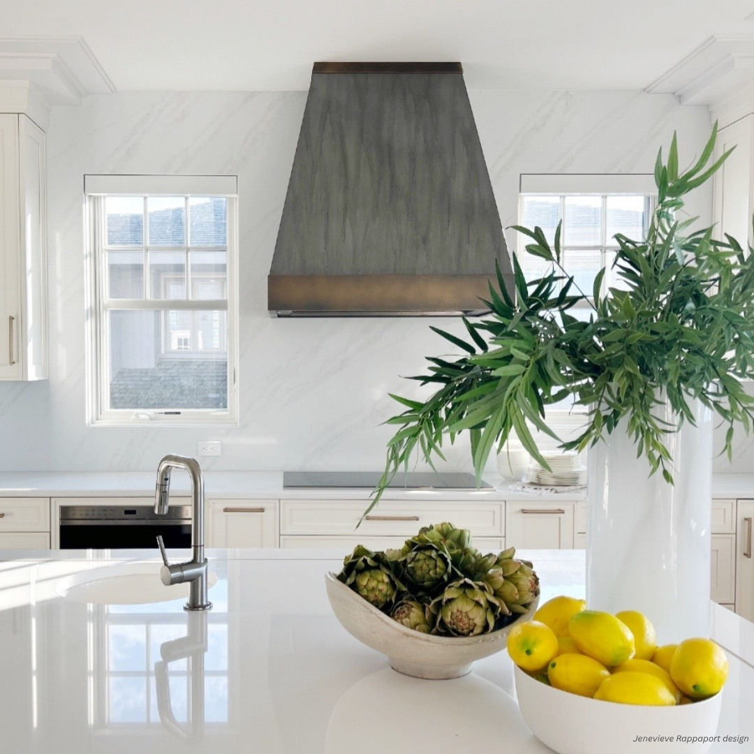 Vail gallery1 features a modern white kitchen with a stainless steel range hood, two windows, and a marble backsplash. The island displays a vase with green branches and a bowl of lemons and artichokes.