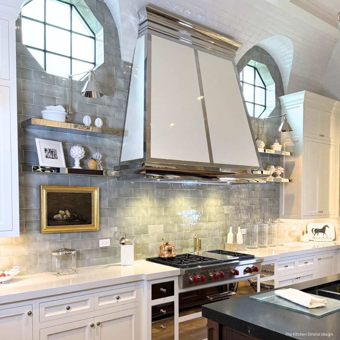 The Vail gallery2 copy features a bright, modern kitchen with glossy gray subway tile, open shelves for decor, a white and chrome range hood, gas stovetop, marble counters, arched windows, white cabinets, and brass accents.