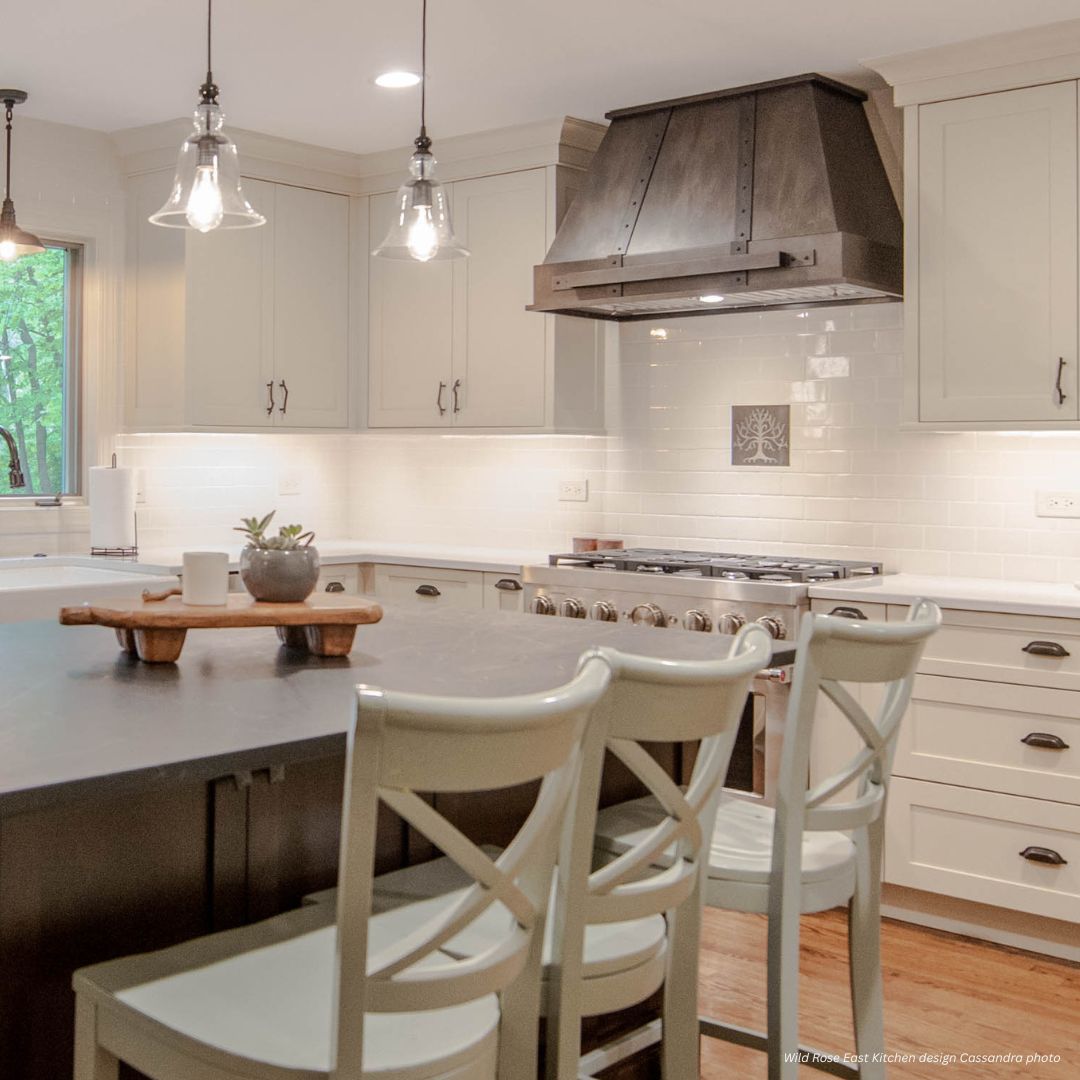 Modern kitchen featuring Vail Gallery3 white cabinets, a dark island with three light barstools, stainless steel appliances, a wood range hood, pendant lights, and a window for natural light.