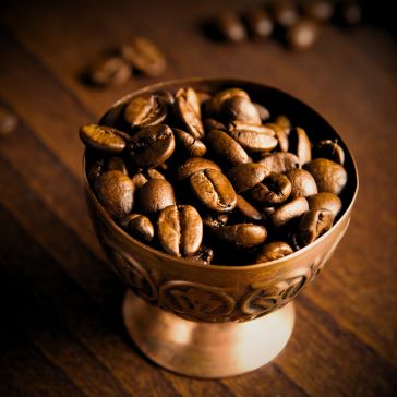 Warm Foundry 1, a brass cup filled with roasted coffee beans, rests on a wooden surface with scattered beans in the background.