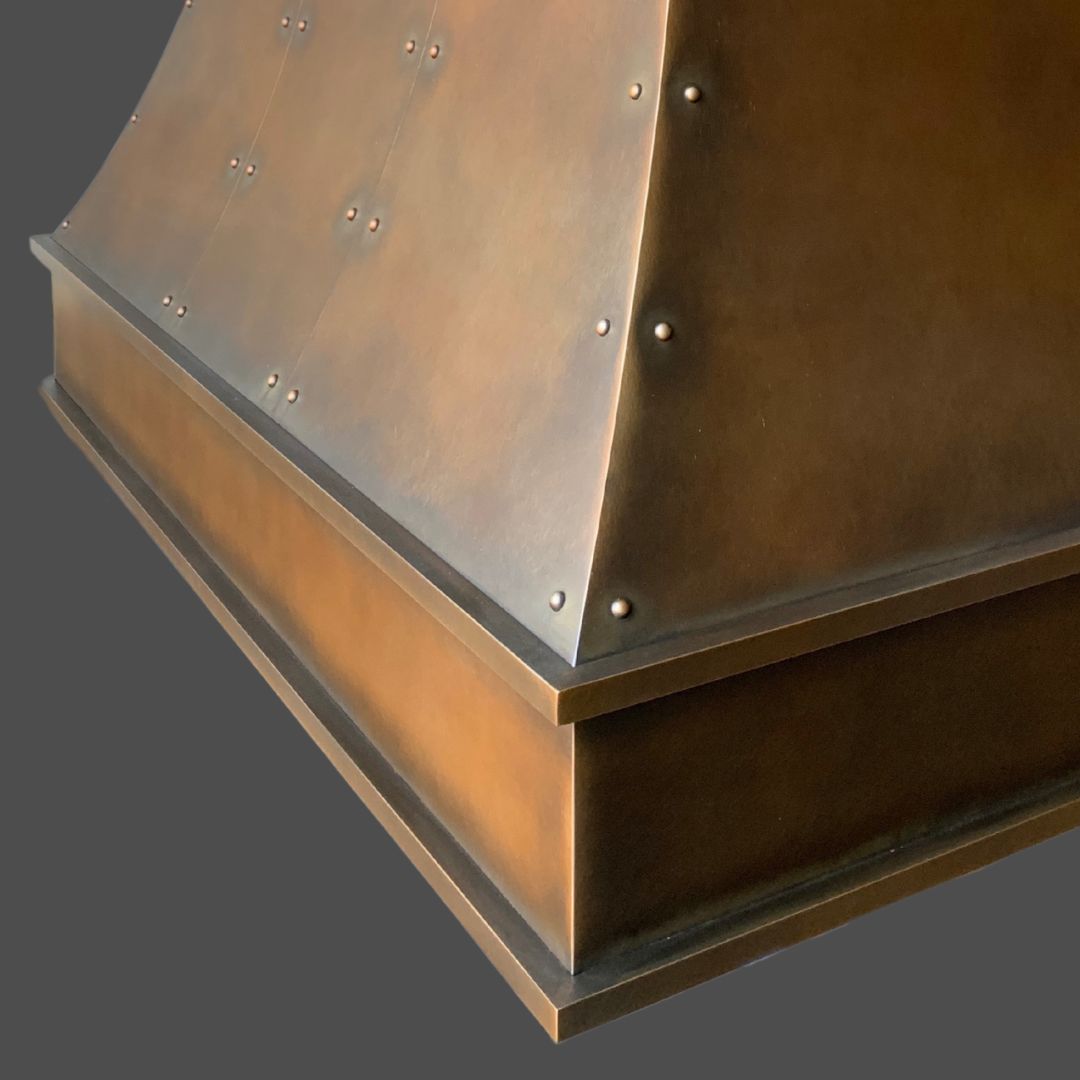 Close-up of Warm Foundry 3, a metallic riveted structure with a bronze or copper finish, featuring a curved surface and angular base, set against a plain gray background.