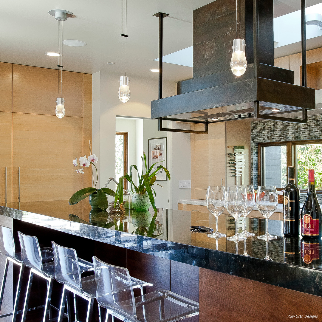The Wash Park gallery2✅ features a modern kitchen with a glossy black island countertop, clear acrylic bar stools, wine glasses, bottles, a potted orchid, hanging lights, wood cabinets, and stainless steel appliances.