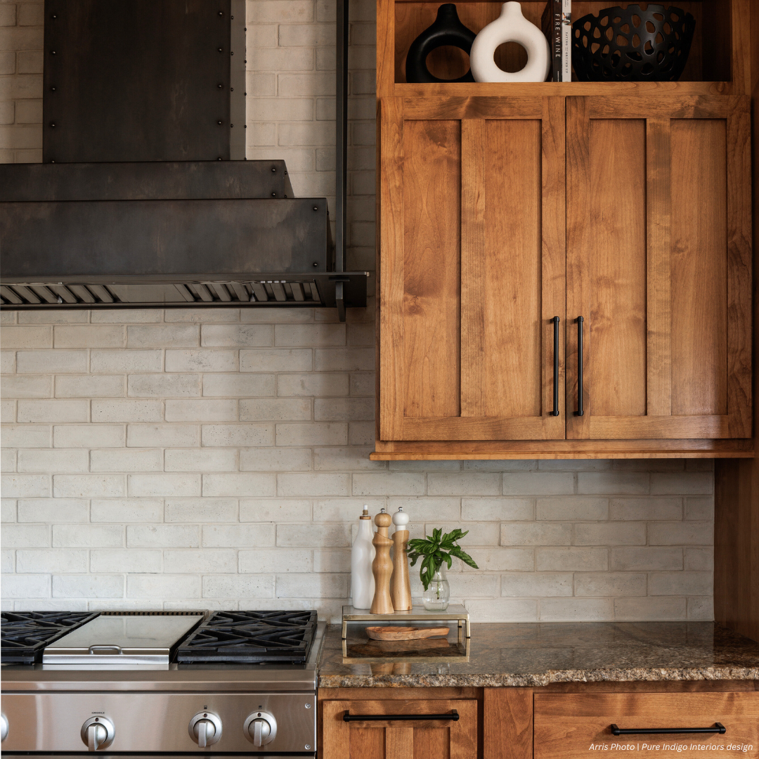 A modern kitchen from Wash Park gallery3 featuring light wood cabinets, a stainless steel gas stove, dark metal range hood, granite countertop with salt and pepper shakers, a small vase with greenery, and a white brick backsplash.