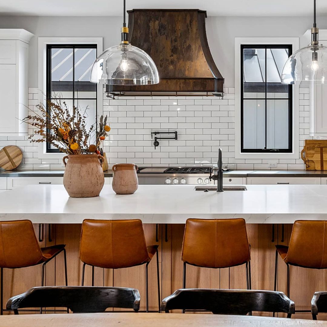 Winter Park gallery2✅ Winter Park gallery2 showcases a modern kitchen with a large island, brown leather chairs, white countertops, dark metal range hood, white subway tile backsplash, two glass pendant lights, and a vase with dried flowers on the island.