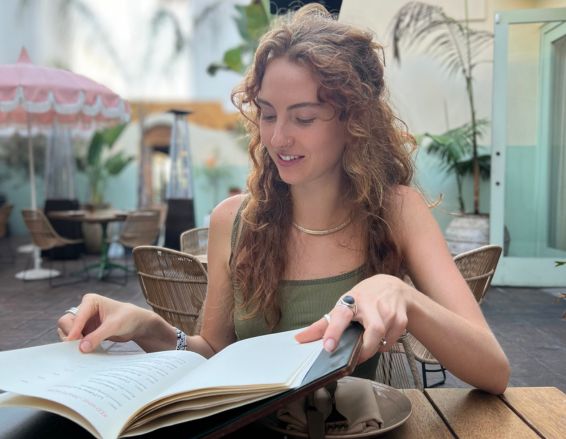 Addison Sasick sits at an outdoor café table, smiling as she looks at an open book. The scene features curly hair, wicker chairs, lush plants, and a pink umbrella in the background.