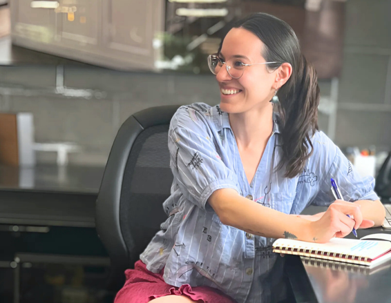 Caroline, a young woman with glasses and a ponytail, smiles as she writes in her notebook at a desk. She wears a light blue shirt and pink skirt, looking happy and engaged.
