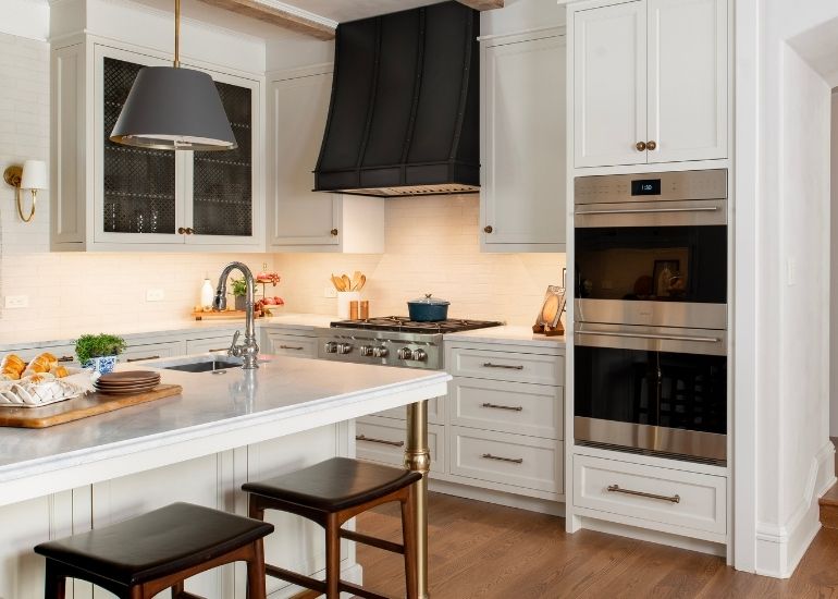 EVG Avon banner The EVG Avon banner showcases a bright, modern kitchen with white cabinets, a large marble island with pastries and dishes, two wooden stools, stainless steel oven, gas stove, black range hood, and wooden floors.