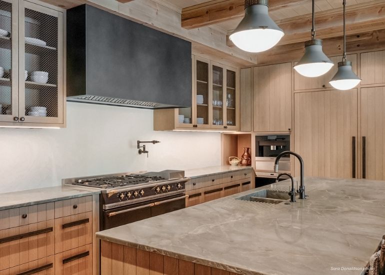 Raw Urth Box Modern kitchen features light wood cabinets, marble countertops, stainless steel appliances, a Raw Urth Box black range hood, pendant lights, open shelving with dishes, and a large island with sink.