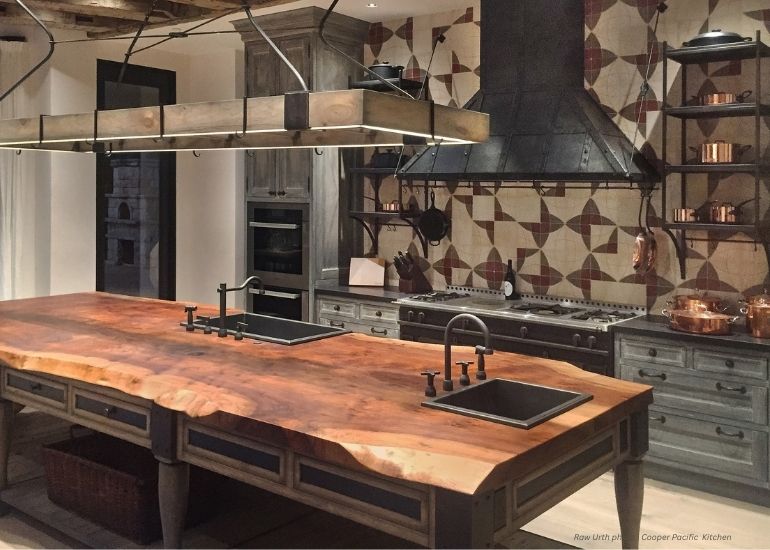 Rustic kitchen features a large wooden island with double sinks, professional stove topped by the Raw Urth Breckenridge black range hood, geometric backsplash, hanging pot racks, and cabinetry along the back wall.
