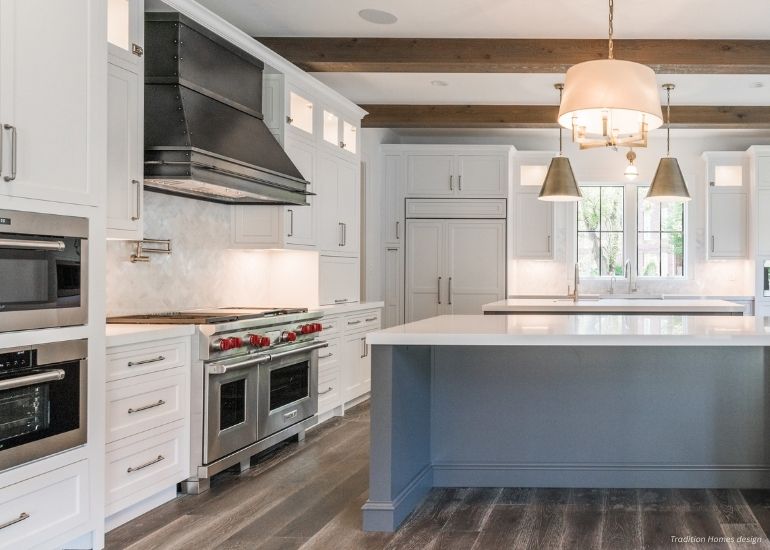 Raw Urth Creede Modern kitchen featuring Raw Urth Creede range hood, white cabinets, stainless steel appliances, a large gray-based island, pendant lighting, wood ceiling beams, and natural light from the window above the sink.