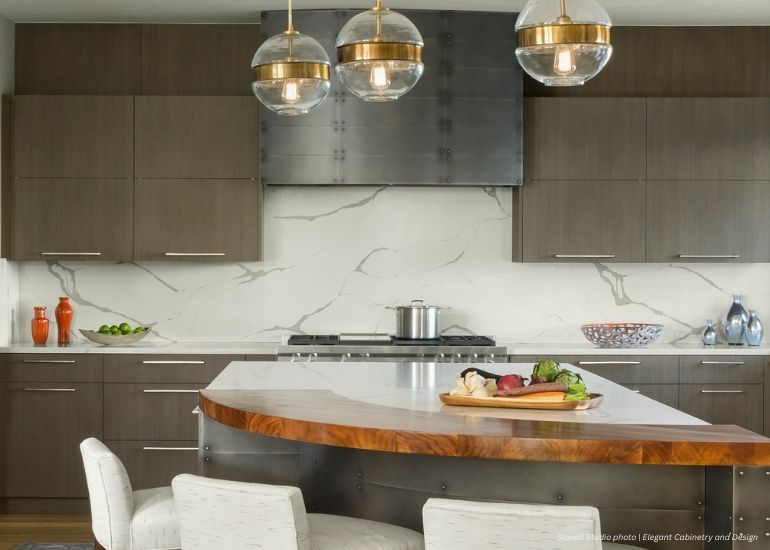Raw Urth Gunnison Modern kitchen featuring dark wood cabinets, white marble backsplash, stainless steel appliances, and a large Raw Urth Gunnison island with a wood countertop. Three pendant lights hang above the island, which holds vegetables and a bowl.