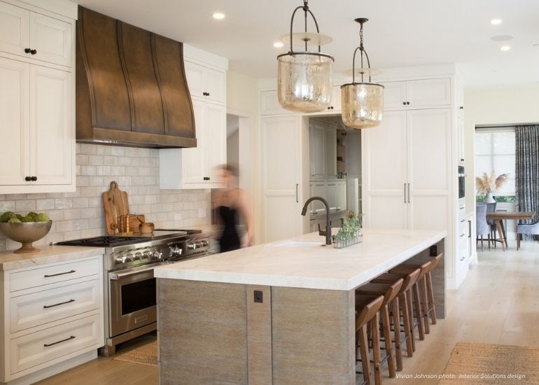 Raw Urth Masonville A modern kitchen with a Raw Urth Masonville metal range hood, large island, wooden stools, white cabinets, stainless steel appliances, pendant lights, and subway tile backsplash. A blurred person walks near the stove.